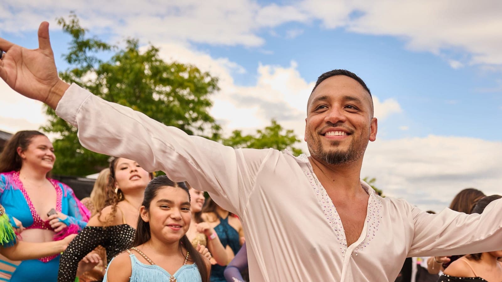 A smiling dancer with arms spread wide leads a line of costumed dancers at Latin Sparks, Ottawa — Latin Sparks × Karmaflow customer story